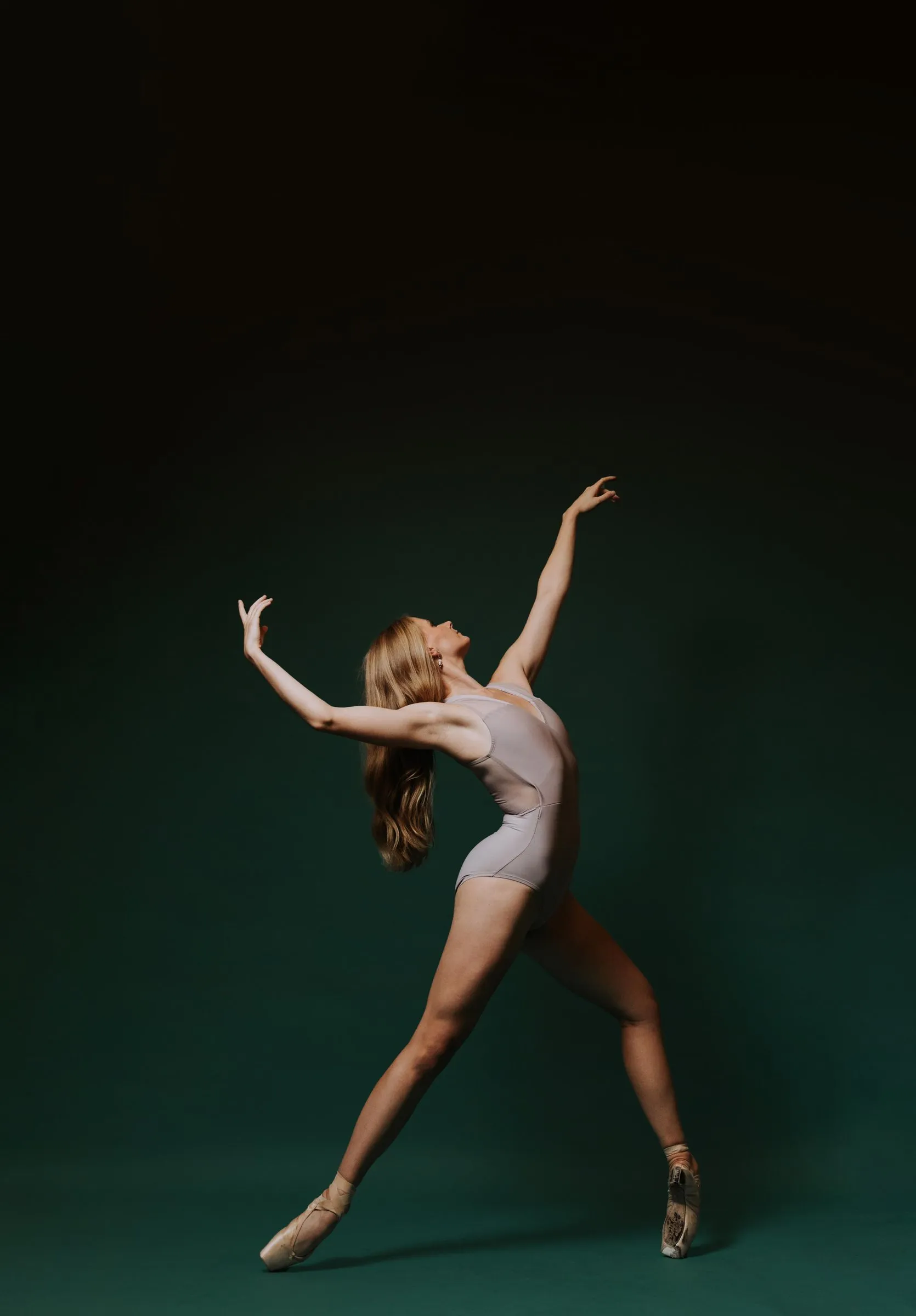 A dancer in a pale leotard arching back with arms open against teal