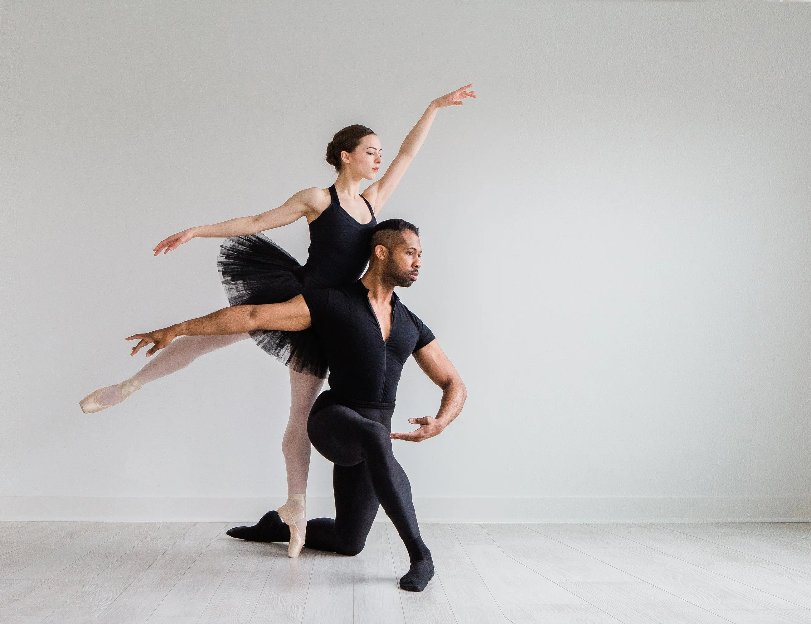 Two dancers in black in a white studio, one in arabesque over the other’s kneeling partner